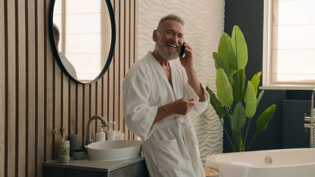 Happy smiling Caucasian retired elderly man talk smartphone friendly call make distant order speak cellphone senior mature old 60s male in bathroom in bathrobe talking mobile phone morning hygieneの写真素材