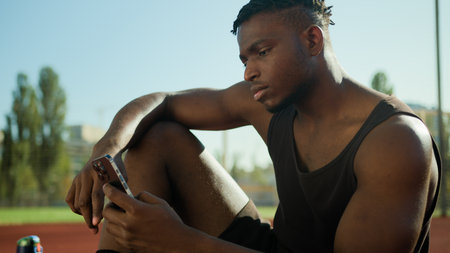 African American man guy sportsman scrolling smartphone sport and technology in city male use mobile phone having break outdoors on sports court athlete using cellphone outside relaxing post-workoutの写真素材