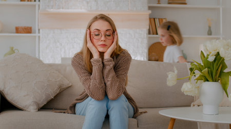 Tired upset Caucasian mother sitting on sofa at home sad frustrated stressed exhausted fatigued mom suffer headache with active noise difficult kid child baby girl daughter running around playingの写真素材