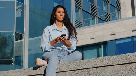 Pensive stressed young woman girl with mobile phone sits in city near building thinks about problem...