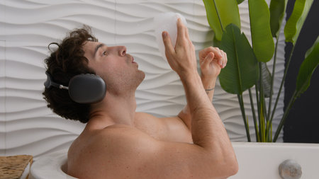 Calm tranquil Caucasian naked relaxed guy sitting in bathroom resting man relax in foamy bathtub blow foam enjoy audio in earphones male relaxing at home bath hotel listening music song in headphonesの写真素材