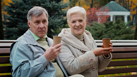Carefree elderly grandparents drinking hot coffee tea outdoors senior aged couple man woman...