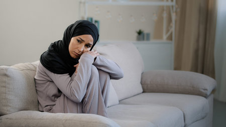Anxious sad young woman in black hijab sitting on couch in living room suffering from psychological...