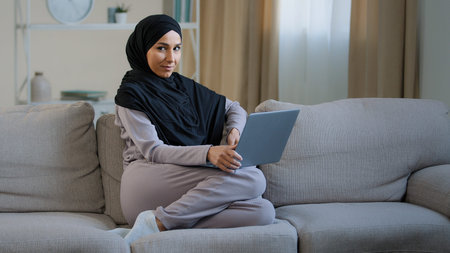 Attractive young muslim woman wear hijab sit on cozy sofa in living room adorable energetic...