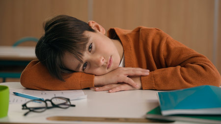 Caucasian bored schoolboy sleepy at school elementary education study learning tired exhausted sad upset fatigued child boy kid pupil schoolchild lying at table desk lesson class relax health problemの写真素材