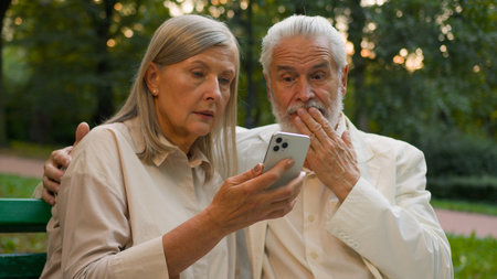 Worried aged 70s Caucasian family retired couple senior man woman using mobile phone device scrolling internet website users customers talking call lost connection sit bench outdoors city nature parkの写真素材