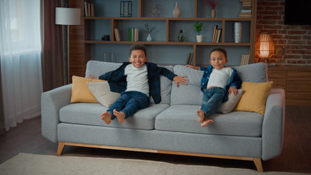 Happy family two African American ethnic little boys children siblings kids running in living room carefree funny play jumping on soft sofa laugh jump on cozy couch relocation at home playing gameの写真素材