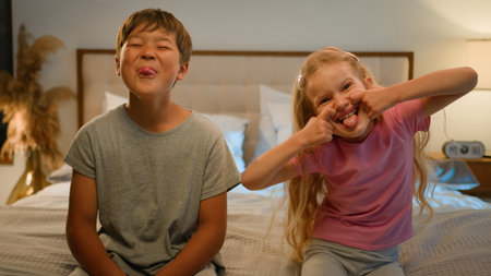 Funny happy Caucasian children kids boy girl brother sister siblings son daughter friends teasing camera at home grimacing playing together fooling around making faces showing tongue joking in bedroomの写真素材
