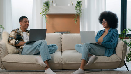 Happy couple relax at couch at home with laptop internet overuse freelancers African American man with woman smiling positive at sofa using typing computers addict pc devices gadgets online gaming appの写真素材