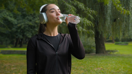 Happy beautiful Caucasian woman fitness girl smile in city park sport outdoors female listening music headphones relax drink water healthy lifestyle refreshment thirst supply drinking aqua hydrationの写真素材