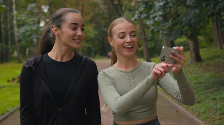 Two happy Caucasian sport girls bloggers influencers women using mobile phone smartphone call record video blog vlog healthy lifestyle workout in city active smiling female sportswomen walking in parkの写真素材