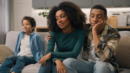 Sad African American family desperate parents hug unhappy mother woman female dissatisfied dad man male sit couch ignore misbehaves little child loud shouting boy active kid son jumping sofa room homeの写真素材