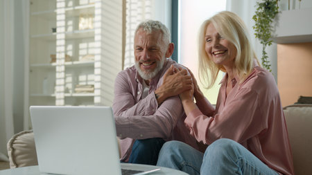 Happy senior family Caucasian couple elderly woman man talking online video call female male using laptop computer pc mature husband wife laugh cuddle hug talk internet virtual chat indoor living roomの写真素材