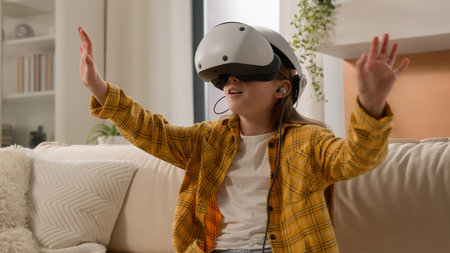 Amazed modern little Caucasian child girl daughter play video game online VR virtual reality glasses headset device. Schoolgirl kid having fun alone 3D cyberspace using innovative technology home roomの写真素材