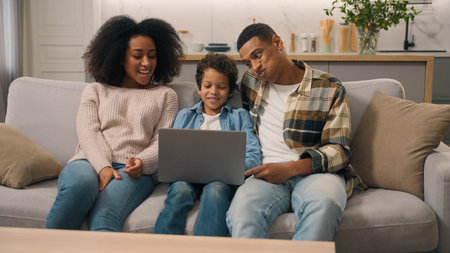 Happy African American family using modern laptop. Mother mom woman female dad father man male help little boy child kid son study learning online education notebook homework talk discuss indoors homeの写真素材