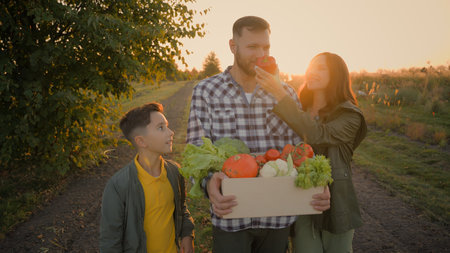 Happy Caucasian family return after farming work harvesting man male guy father carrying box crate vegetables little child son kid boy stroll woman female mother walking field enjoy sniff organic foodの写真素材