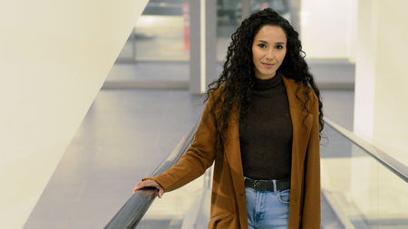 Young beautiful brunette woman riding on escalator indoors smiling looking at camera. Girl in...