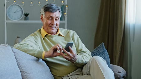 Caucasian elderly adult man sitting on sofa at home with gadget using online app on smartphone makes online order old grandfather resting reading news in mobile phone browsing scrolling websitesの写真素材