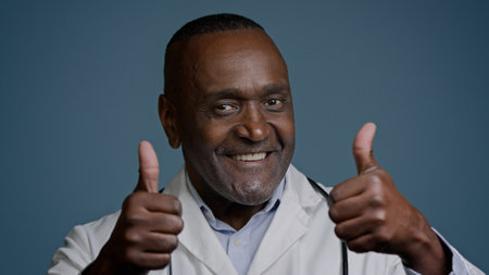 Portrait smiling African American man doctor in white coat showing thumbs up in studio blue background looking at camera gesture approval recommendation medical services health worker show like signの写真素材