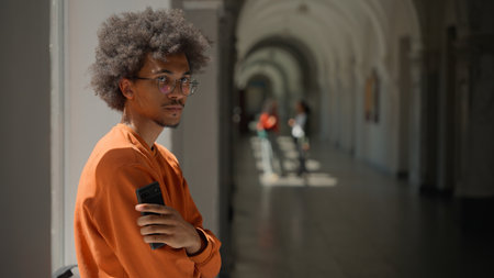 African American student guy using mobile phone in university hall corridor near window looking holding smartphone app chatting internet education study break man male pensive serious with cellphoneの写真素材