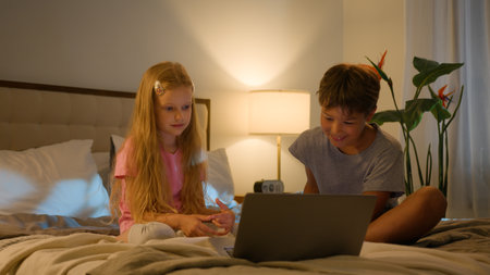 Happy Caucasian kids little siblings smiling family brother boy son daughter girl children watching online video enjoying cartoon movie using modern tech pc laptop computer bed indoors evening bedroomの写真素材