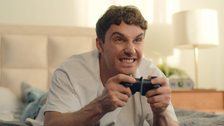 Playful Caucasian European young man lying on bed playing console holding joystick video games fun...