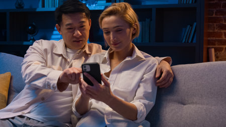 Family multiracial couple Asian man and Caucasian woman at night home buying goods in Internet store mobile phone shopping online with smartphone browsing together talking discuss order food deliveryの写真素材