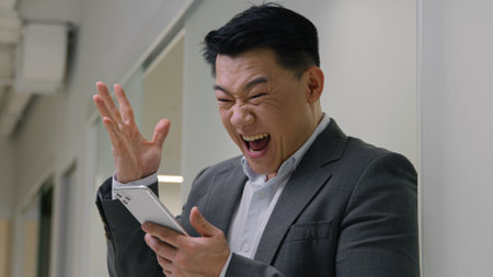 Excited guy in hallway office business man middle-aged entrepreneur Asian chinese korean multiethnic businessman winning mobile phone bet app online lottery male winner win scream shout achievementの写真素材