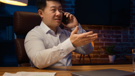 Happy Asian man worker manager executive boss entrepreneur at night office talking mobile phone at desk. Korean businessman Chinese company CEO Japanese investor talk smartphone call at late eveningの写真素材