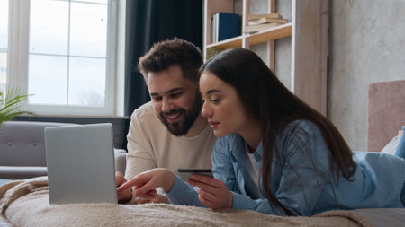 Caucasian couple family lying together on bed use credit bank card enter code in laptop computer banking app online payment smiling woman and man shopping internet order pay delivery at home bedroomの写真素材