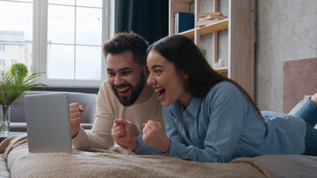 Excited Caucasian married couple lying on bed at home win laptop achievement read amazing news online bet victory celebrate success happy woman wife and man husband scream shout good result winningの写真素材