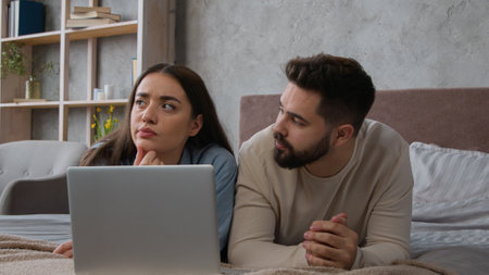 Caucasian couple married wife husband lying on bed family talking discuss shopping online together use laptop internet order young woman man clients looking at computer choosing home goods for bedroomの写真素材