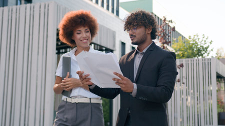 Indian Arabian male man businessman holding papers documents corporate discuss result plans with African American woman colleague businesswoman walking downtown in city multiracial business peopleの写真素材