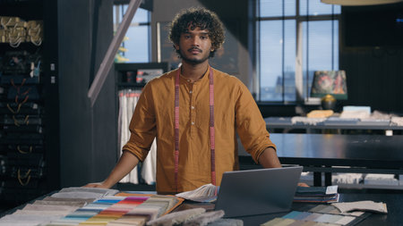 Indian Arabian man happy smiling creative designer fashion clothing tailor posing crossed hands in modern studio textile workshop with fabric color swatches for interior design project small businessの写真素材