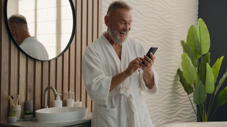 Old Caucasian man senior mature retired male in bathrobe in bath bathroom at morning looking mobile phone reading shocked good news amazed wonder wow reaction surprise smartphone sms message smilingの写真素材