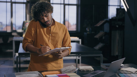 Arabian Indian male business man working busy interior designer talking with client online discuss on mobile phone fabric tissue samples write in notepad textile order in workshop modern design studioの写真素材