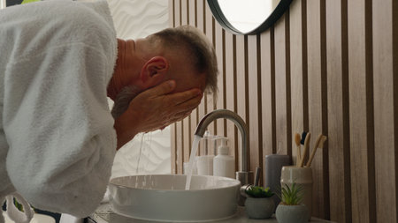 Old Caucasian gray-haired man washing face with cold warm hot water middle-aged senior male retired mature pensioner wash facial hygiene moisturize refresh in morning bath sink skin care refreshingの写真素材