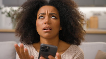 Worried frustrated African American ethnic woman on couch holding mobile phone in hands receive message bad news upset stressed anxious shocked girl female lady problem cell smartphone failure crashの写真素材