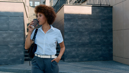 Moving shot happy confident African American business woman girl lady ethnic female businesswoman walking in city walk going move holding cup take away coffee tea drink urban building office skylineの写真素材