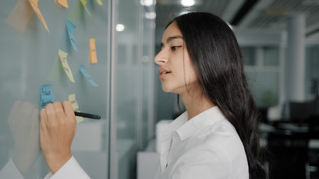 Thoughtful young businesswoman arabian hispanic creative designer come to note board in office read...