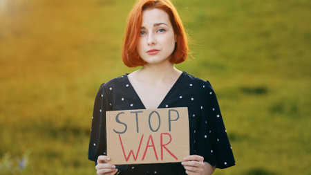 Unhappy caucasian young redhead woman activist hold demonstrating banner with inscription stop war sad pretty girl stand outdoors in field show cardboard protests against military conflict in Ukraineの写真素材