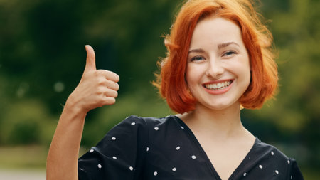 Stylish young pretty girl attractive caucasian redhead teen woman girlfriend cheerful smiling friendly looks at camera puts thumb up gesture recommends best choice shows everything fine stands outsideの写真素材
