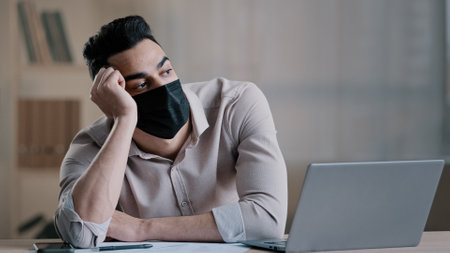 Bored sad hispanic worker young business man in medical mask tired unmotivated employee disinterested in dull distant work on computer lazy male arab guy feeling lack of motivation boredom in officeの写真素材