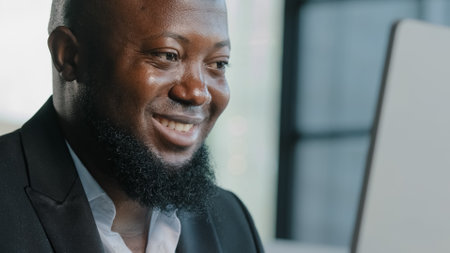 Smiling african businessman adult professional employee web designer chatting in networks using wifi internet at office searching digital information on laptop working online watching business webinarの写真素材