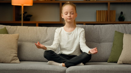 Little cute caucasian girl young healthy child blonde kid preschooler meditate sit on sofa at home calm mindful peaceful daughter do yoga breathing exercise relax alone with closed eyes in lotus poseの写真素材