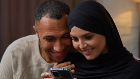 African american man join to muslim woman sitting with mobile phone multiracial diverse ethnic couple holding smartphone watching funny video on 5g internet buying home goods online modern tech usageの写真素材
