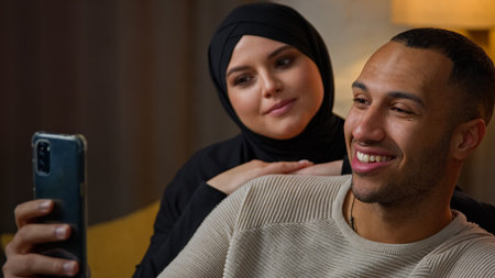 Family ethnic multiracial african american man and arabian muslim woman chatting online talking use mobile phone webcam app internet video call at home. Couple speaking virtual meeting by smartphoneの写真素材