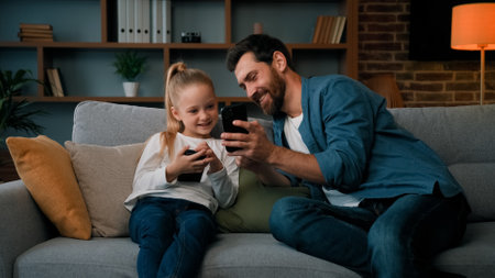 Caucasian man and little kid girl sit together at home hold modern phone browsing media content play games online adult dad teach child daughter use mobile apps showing funny video on wireless deviceの写真素材