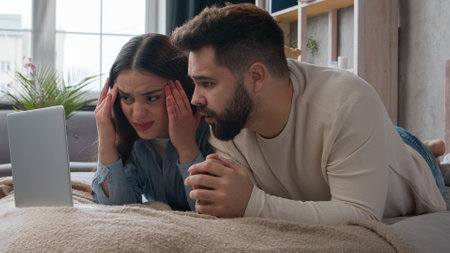 Caucasian family couple upset disappointed read news result refusal email with failure shocked woman and man homeowners on bed use laptop computer watch movie in bedroomの写真素材