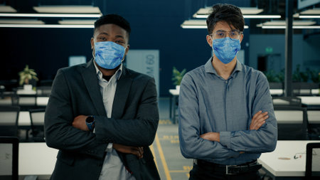 Business people two confident colleagues office workers african indian ethnic men stand with crossed arms in face medical mask. Covid-19 corporate measures prevent disease grippe coronavirus symptomsの写真素材